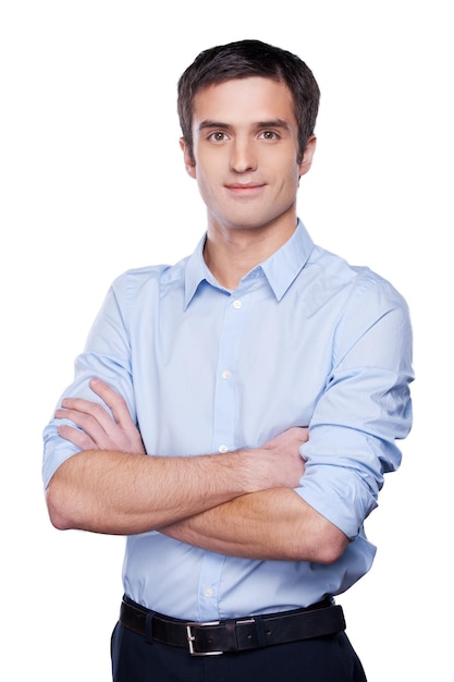 Joven y exitoso. Apuesto joven con camisa azul mirando a la cámara y manteniendo los brazos cruzados mientras está de pie aislado en blanco