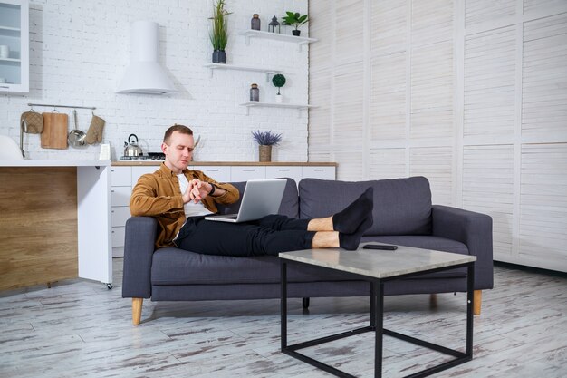 Un joven exitoso está sentado en su casa en el sofá con una computadora portátil y está trabajando. Trabajo remoto durante cuarentena.