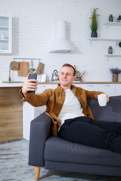 Un joven exitoso está sentado en su casa en el sofá con un teléfono y hablando por un enlace de video. Trabajo remoto durante cuarentena.