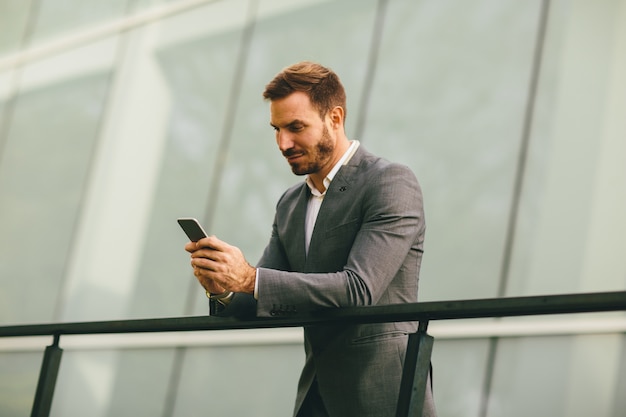 Joven exitoso hombre de negocios con traje gris y sosteniendo su teléfono inteligente mientras está de pie cerca de la moderna