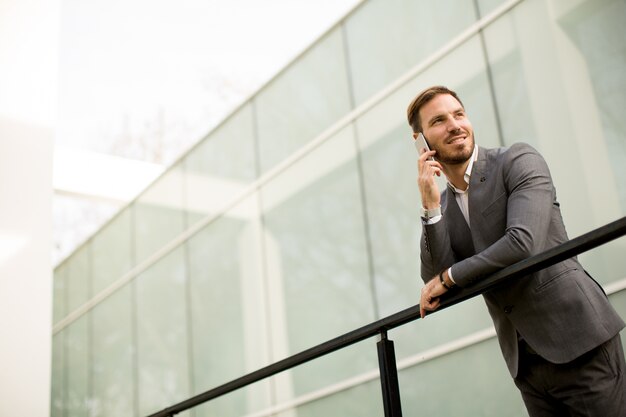 Foto joven exitoso hombre de negocios con traje gris y sosteniendo su teléfono inteligente mientras está de pie cerca de la moderna