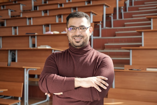 Joven y exitoso profesor de la universidad cruzando los brazos por el pecho mientras está de pie frente a la cámara contra los escritorios en la sala de conferencias