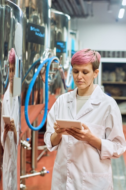 Joven experta en cervecería de bata blanca mirando a través de datos en tableta digital mientras está de pie junto a nuevos equipos para la producción de cerveza en la planta