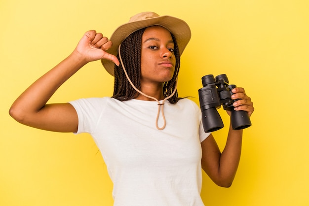 Joven exploradora afroamericana sosteniendo un binoculares aislado sobre fondo amarillo se siente orgullosa y segura de sí misma, ejemplo a seguir.