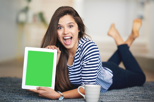 Una joven extasiada tirada en el suelo en casa sosteniendo una tableta digital con una pantalla de croma