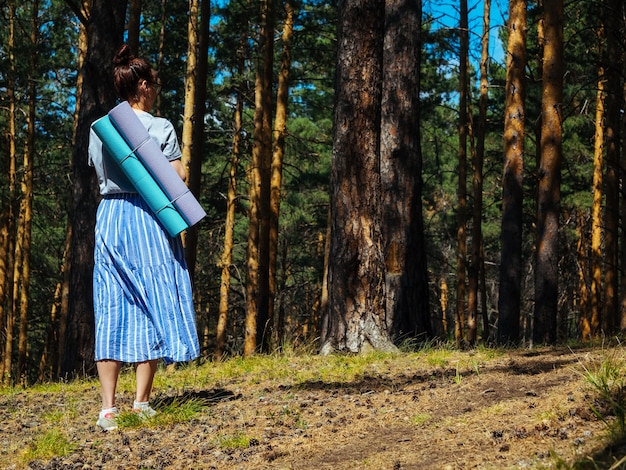 Una joven con una falda ligera camina por el bosque y lleva colchonetas de yoga a la espalda, copyspace,