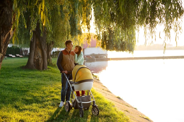 Una joven familia amorosa camina por el lago con una carriola sonriente pareja de padres con un cochecito de bebé en