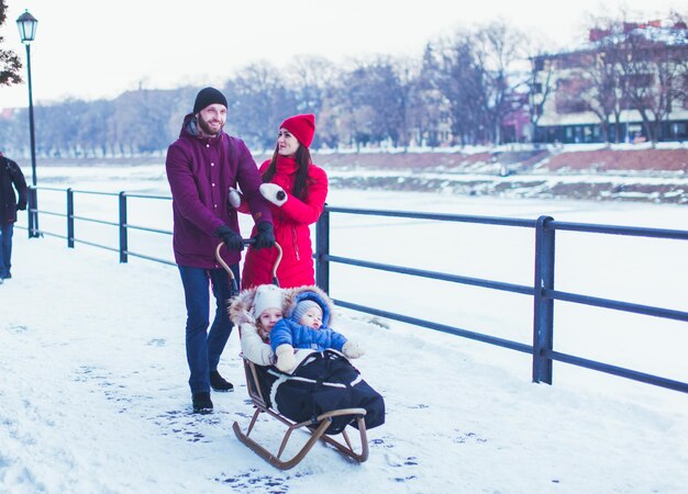 Joven familia atractiva con ropa de invierno cálida y brillante caminando con su adorable hija pequeña y su lindo hijo. Encantadores niños sentados en trineo, empujados por su padre