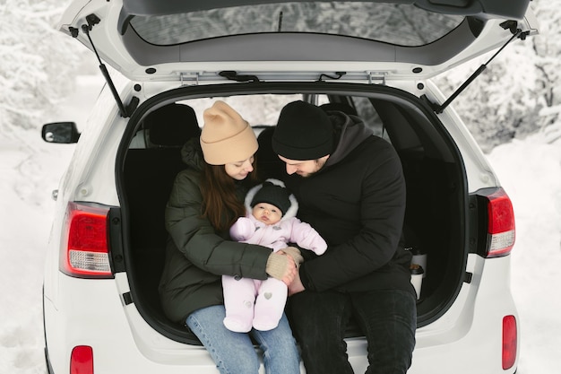 Una joven familia feliz, un hombre, una mujer y un bebé, están sentados en el maletero de un coche en invierno