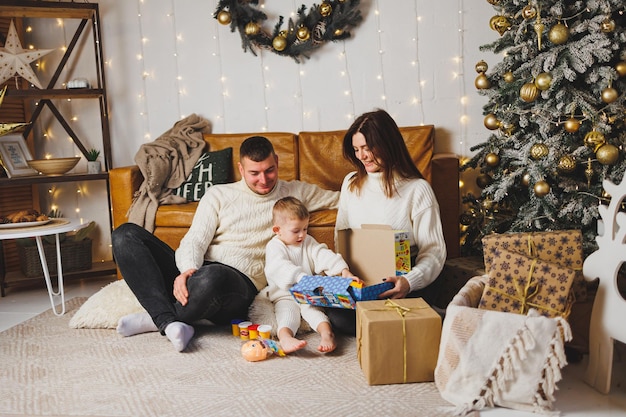 Una joven familia feliz con un niño pequeño celebra el Año Nuevo en casa cerca del árbol de Navidad Navidad con la familia Año Nuevo para celebrar