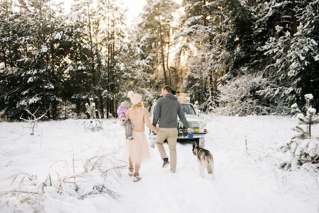 Una joven familia feliz con un niño pequeño se está preparando para la Navidad, caminando con un perro husky en el fondo de un automóvil retro, en el techo de un árbol de Navidad y regalos en el bosque nevado de invierno.