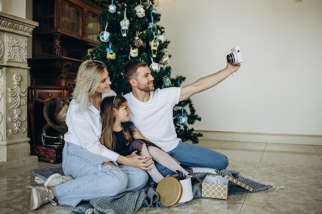 Joven familia feliz de tres tomando una foto de sí mismos en la sala de estar en la víspera de Navidad Familia caucásica con perro sentado junto al árbol de Navidad en casa tomando selfie con teléfono móvil