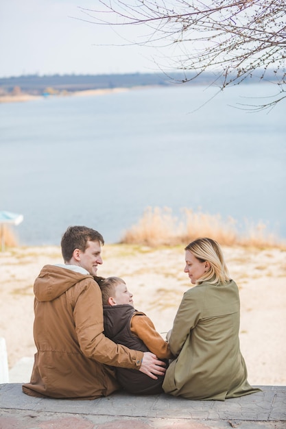 Joven familia hermosa sentada en la playa con vista al lago. Otoño