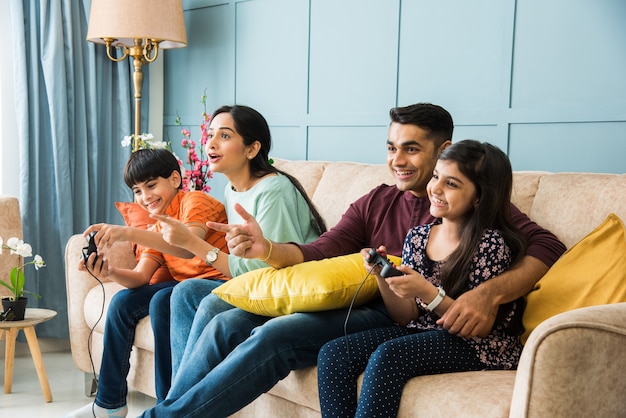 Joven familia india de cuatro jugando videojuegos con controlador o joystick mientras está sentado en el sofá