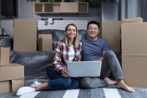 Joven familia multirracial feliz en un apartamento nuevo con una laptop asiática y una mujer mirando la cámara y sonriendo charlando con amigos en línea mostrando un nuevo hogar