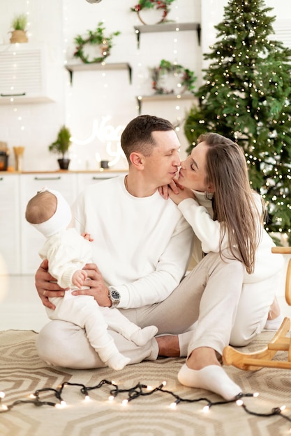 una joven familia con un niño pequeño está disfrutando del año nuevo en el árbol de Navidad en la cocina o en una casa brillante sonriendo besándose y abrazándose felicitándose unos a otros celebra la fiesta