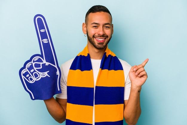 Joven fanático de los deportes hispano aislado de fondo azul sonriendo y señalando a un lado mostrando algo en el espacio en blanco