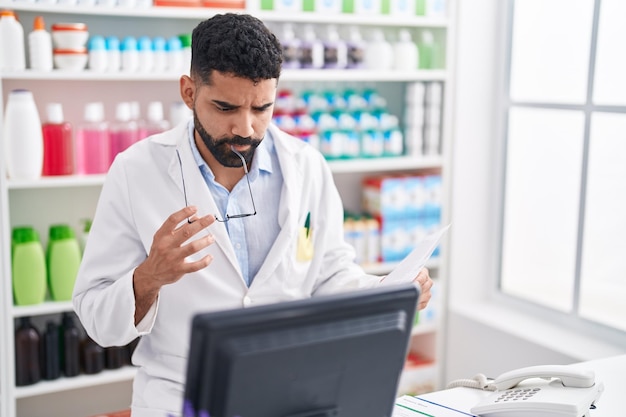 Joven farmacéutico árabe leyendo un documento con expresión de duda en la farmacia