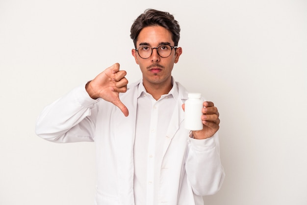 Joven farmacéutico hombre de raza mixta sosteniendo píldoras aisladas sobre fondo blanco mostrando un gesto de aversión, pulgares hacia abajo. Concepto de desacuerdo.