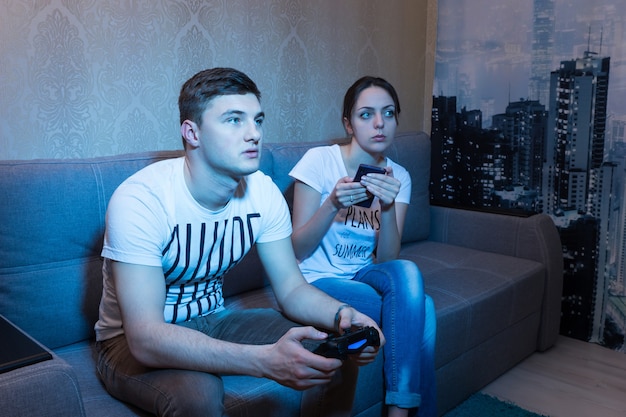 Joven fascinado jugando un videojuego con su novia sentada cerca de él en el sofá y monitoreando el proceso de su juego en casa en un ambiente relajado.