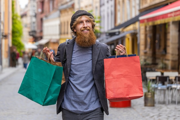 Joven feliz adicto a las compras consumidor después de la venta de compras con bolsas llenas caminando por la calle de la ciudad