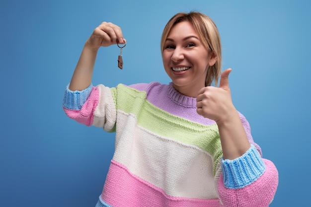 Una joven feliz y afortunada con un suéter informal sosteniendo un llavero de su nuevo apartamento de fondo azul