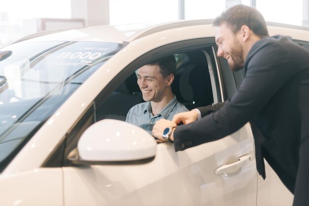 Un joven feliz al volante de un auto nuevo en un concesionario de automóviles Un vendedor de autos profesional con traje de negocios habla sobre un modelo de auto nuevo