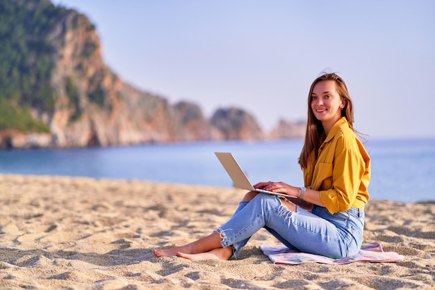 Joven, feliz, alegre, satisfecha, milenaria, independiente, niña, usar la computadora portátil, en la playa, por, mar, sueño, oficina, trabajo, concepto