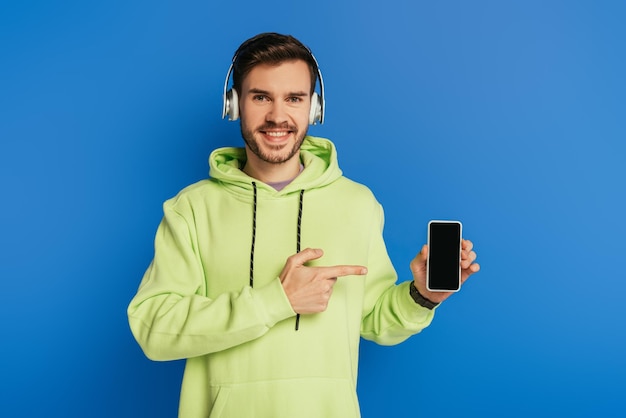 Joven feliz con auriculares inalámbricos apuntando con el dedo al teléfono inteligente con pantalla en blanco aislada