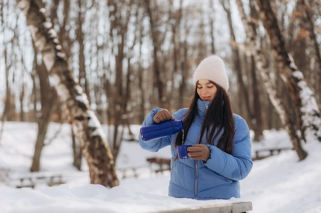 Una joven feliz bebe té caliente de una taza termo durante un paseo por el bosque invernal