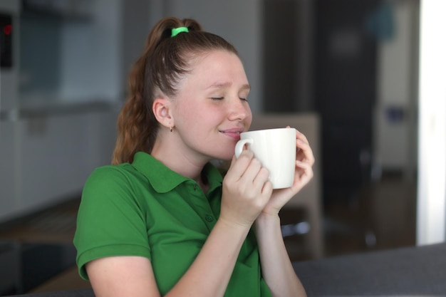 Una joven feliz bebiendo una taza de té soñando con una chica sentada en la sala de estar con una taza de café caliente