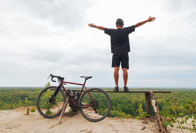 Foto joven feliz con una bicicleta se encuentra en un acantilado y disfruta de la vista con los brazos extendidos.