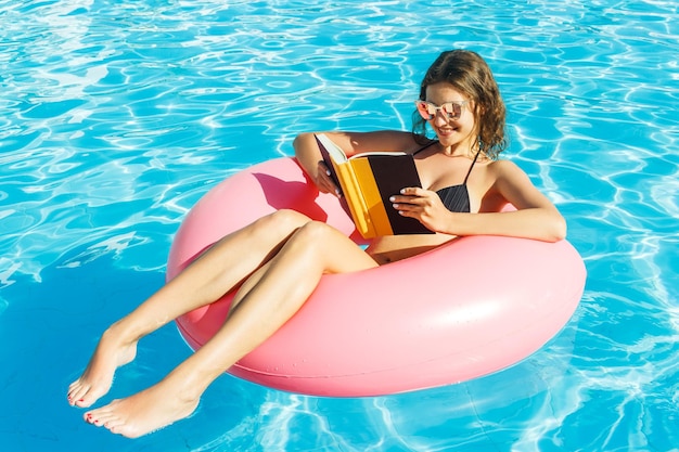 Una joven feliz en bikini está nadando en la piscina y leyendo un libro con un círculo rosa