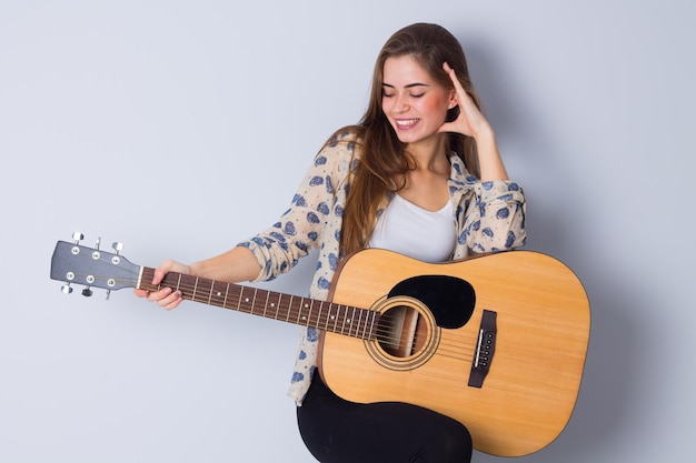 Una joven feliz con blusa beige y pantalones negros sosteniendo una guitarra con fondo gris en el estudio