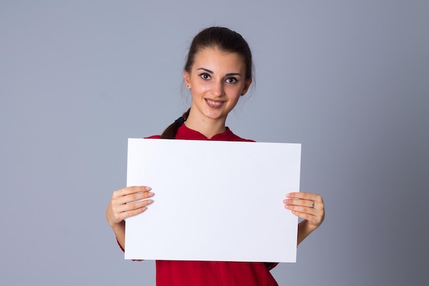 Una joven feliz con blusa roja sosteniendo una hoja blanca de papel con fondo gris en el estudio