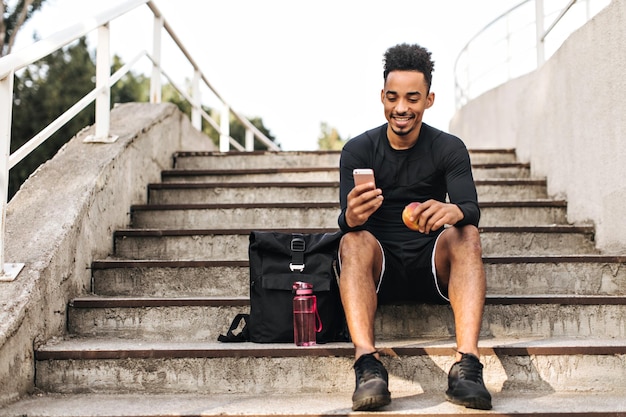 Joven, feliz, brunet, de piel oscura, con pantalones cortos negros y una elegante camiseta deportiva, se sienta en las escaleras afuera, mira la pantalla del teléfono y sostiene la manzana.