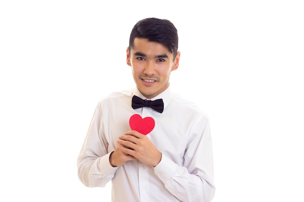 Joven feliz con cabello negro en camiseta blanca con corbatín negro sosteniendo un corazón de papel en el estudio