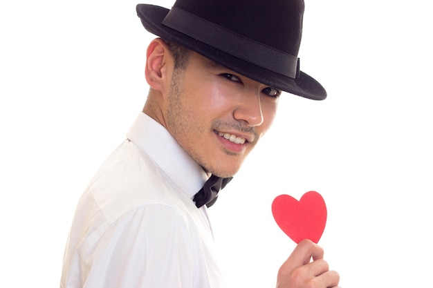 Joven feliz con cabello oscuro en camiseta blanca con pajarita y sombrero negro sosteniendo un corazón de papel rojo sobre fondo blanco en el estudio.