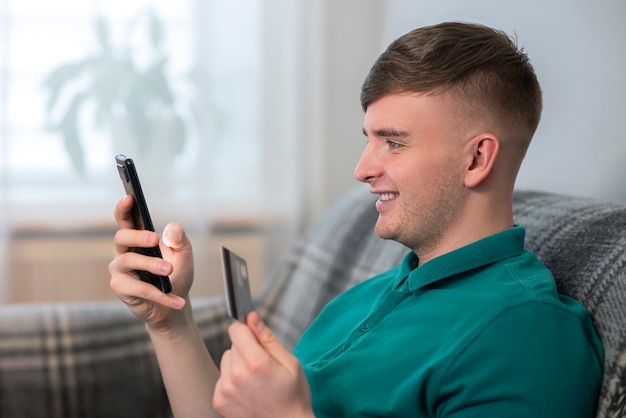 Joven feliz, chico positivo está sosteniendo una tarjeta bancaria de crédito y un teléfono móvil celular en la mano