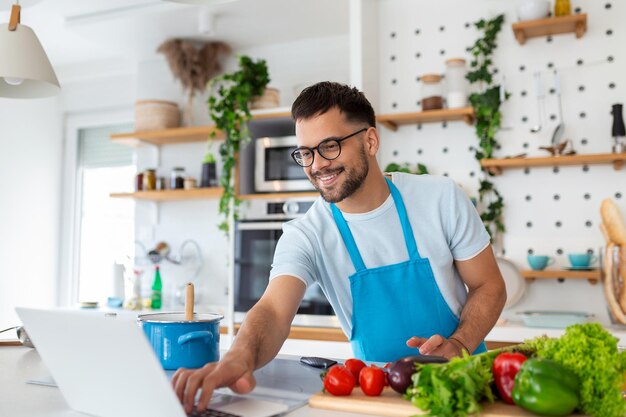 Un joven feliz cocinando una cena saludable en casa está siguiendo un tutorial de video en la computadora portátil