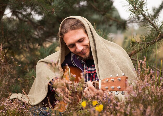 Un joven feliz cubierto de tela escocesa tocando guitarra acústica y cantando. Hombre en el bosque de pinos con prado de flores de brezo. Cerrar, copiar espacio.