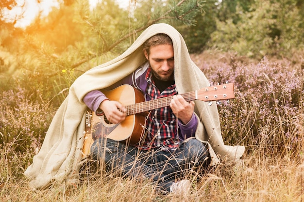 Un joven feliz cubierto de tela escocesa tocando guitarra acústica y cantando. Hombre en el bosque de pinos con prado de flores de brezo. Cerrar, copiar espacio.