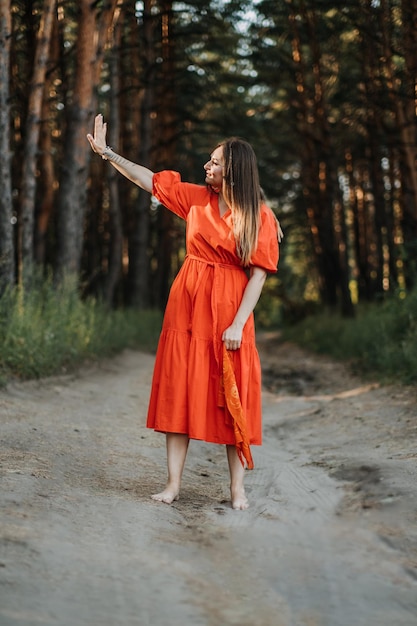 Joven feliz descalza vestida de rojo con la mano levantada bailando en un bosque de pinos en el día de verano