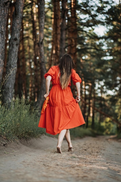 Joven feliz descalza vestida de rojo con la mano levantada bailando en un bosque de pinos en el día de verano