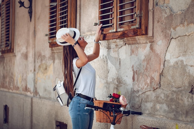 Una joven feliz disfruta de un día soleado de verano, mirando a la ventana de la casa vieja, al lado de la bicicleta con una cesta de flores.