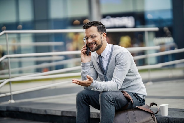 Un joven feliz y elegante está sentado en las escaleras al aire libre y hablando por teléfono inteligente