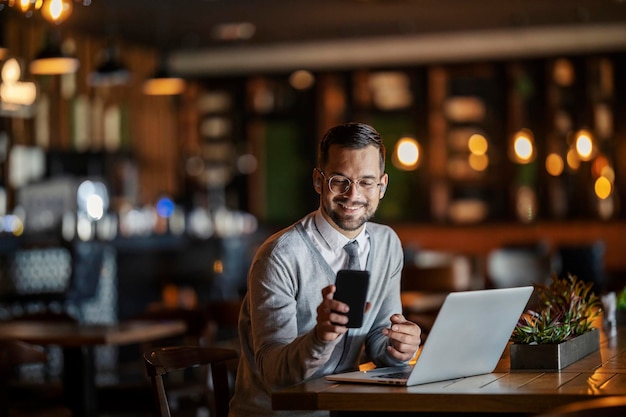 Un joven feliz y elegante hombre de negocios en elegante casual está sentado en una cafetería con su portátil