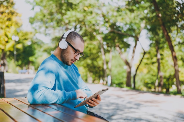 Joven feliz, empresario o estudiante con gafas de camisa azul casual sentado en la mesa con auriculares, tablet pc en el parque de la ciudad, escuchar música, descansar al aire libre en la naturaleza verde. Concepto de ocio de estilo de vida.