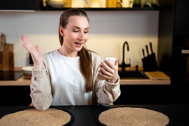 Una joven feliz está parada en casa en la cocina con su teléfono inteligente jugando juegos en línea ganando