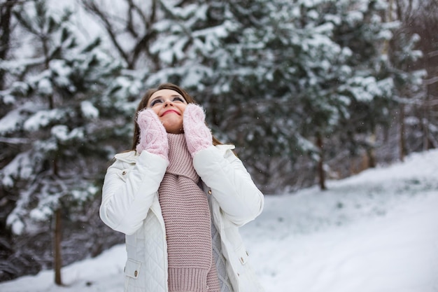 La joven feliz está parada en un parque nevado y mirando hacia arriba. Está vestida con ropa de abrigo con estilo.